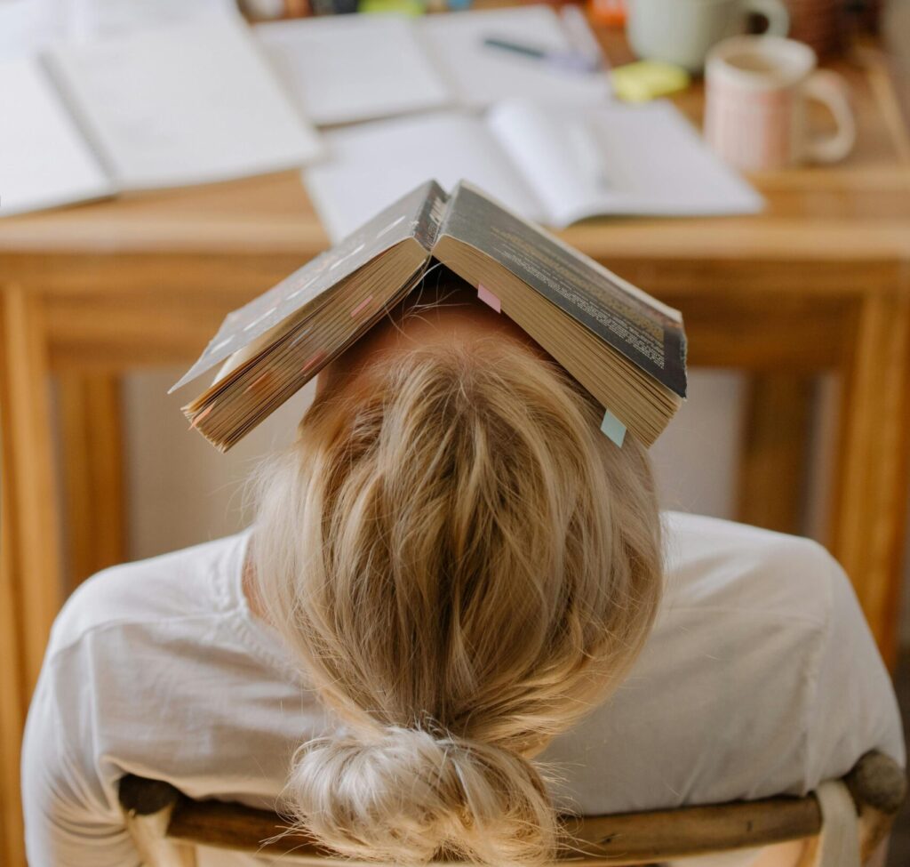 Student feeling stress and exhaustion while studying at a cluttered desk with an open book on their head.