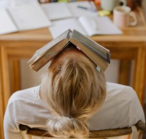 Student feeling stress and exhaustion while studying at a cluttered desk with an open book on their head.