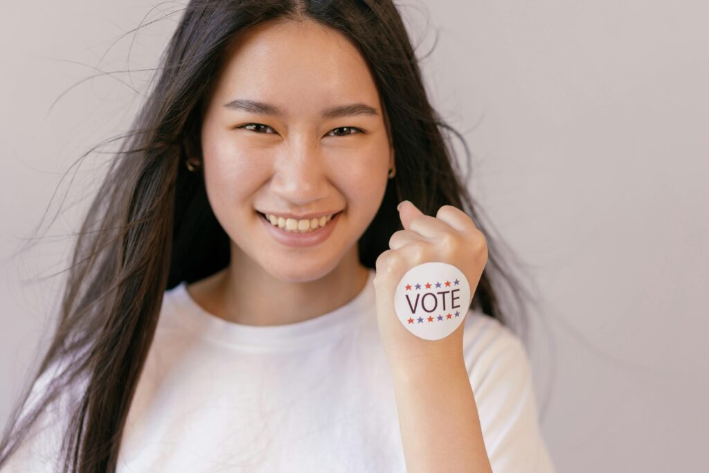 A smiling young woman holds up a vote sticker, promoting the spirit of democracy and participation.